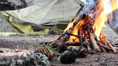 Foto på ett läger. I förgrunden brinner en brasa och i bakgrunden står det ett grönt tält.